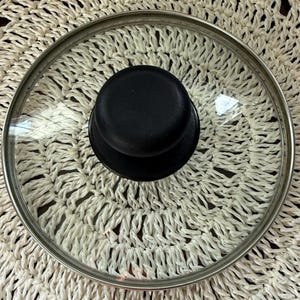 May include: A circular pot lid featuring a clear tempered glass surface, a polished stainless steel rim, and a solid black heat-resistant knob handle centered on top. The lid is designed for standard cookware and provides a clear view of contents.