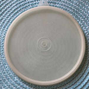May include: A clear, round Tupperware lid with a light beige rim and a small handle. The lid has the Tupperware logo and the number 423-99 printed on it. The lid is made of plastic.