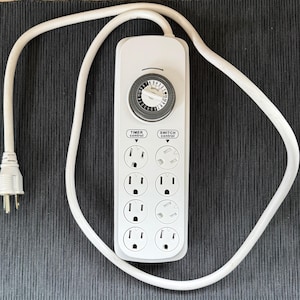 May include: White power strip with a built-in timer and multiple outlets. The timer has a black dial with white markings. The power strip has a white cord and plug. The words "Timer control" and "Switch control" are printed on the power strip.