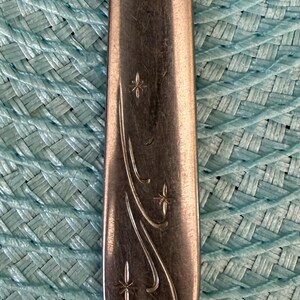 May include: Close-up of a silver spoon with a decorative floral design. The spoon's handle features a curved pattern with star-like accents. The spoon is set against a textured, light blue background.