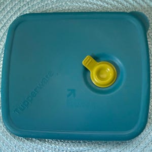 May include: Teal-colored Tupperware lid with a yellow valve. The lid is rectangular with rounded corners and the Tupperware logo printed on the side. A yellow valve is located on the top right side of the lid.