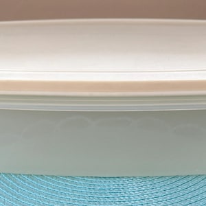 May include: A rectangular, off-white plastic food storage container with a matching lid. The container is light green and sits on a blue placemat. The lid has a slightly darker, beige-colored rim.