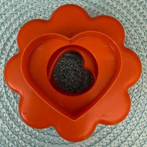 May include: An orange Tupperware cookie cutter set. The set includes a flower-shaped base, a heart-shaped cutter, and a smaller heart-shaped cutter nested inside. The set is made of durable plastic and is designed for easy use and storage.
