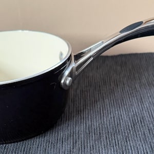 May include: A black saucepan with a silver handle and a cream-colored interior. The handle curves upward and has a black grip. The pan sits on a dark gray surface. The pan is designed for cooking.
