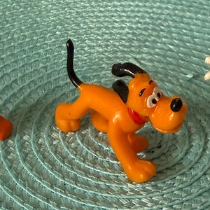 以下が含まれることがあります： ディズニーキャラクターのフィギュア3体：ドナルドダック、プルート、ミッキーマウス。ドナルドダックはセーラー服を着ています。プルートはオレンジと黒です。ミッキーマウスは赤いショートパンツと黄色の靴を履いています。フィギュアは、ターコイズブルーのテクスチャ加工された表面にあります。