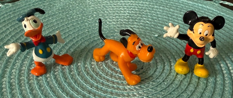 以下が含まれることがあります： ディズニーキャラクターのフィギュア3体：ドナルドダック、プルート、ミッキーマウス。ドナルドダックはセーラー服を着ています。プルートはオレンジと黒です。ミッキーマウスは赤いショートパンツと黄色の靴を履いています。フィギュアは、ターコイズブルーのテクスチャ加工された表面にあります。