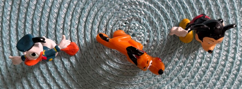 以下が含まれることがあります： ディズニーキャラクターのフィギュア3体：ドナルドダック、プルート、ミッキーマウス。ドナルドダックは青い船乗り帽とジャケットを着ています。プルートはオレンジ色で黒い斑点があります。ミッキーマウスは彼のクラシックな衣装です。