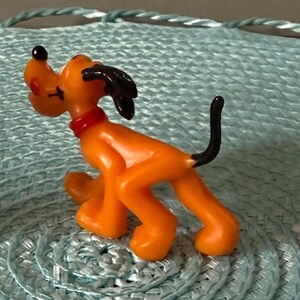 以下が含まれることがあります： ミッキーマウス、プルート、ドナルドダックの3体のディズニーキャラクターのフィギュア。ミッキーマウスは黒、赤、黄色。プルートはオレンジ色で、黒い尻尾と耳。ドナルドダックは白、青、オレンジ色。フィギュアは水色のテクスチャ加工された表面にあります。