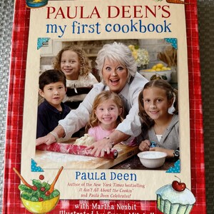 May include: A red and white checkered book cover with the title "PAULA DEEN'S my first cookbook" in white text. The cover features a photo of Paula Deen with four children, all smiling, in a kitchen setting. The book's author and illustrator are listed below the photo.