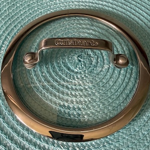 May include: A clear glass Cuisinart pot lid with a stainless steel rim and handle. The handle is curved and has the Cuisinart logo. The lid is circular and sits on a light blue woven surface.