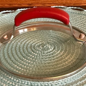 May include: A clear glass lid with a stainless steel rim and a bright red handle. The lid is round and appears to be for a pot or pan. The handle is curved and easy to grip.