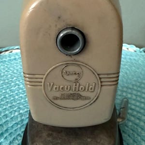 May include: A vintage Apsco Vacu Hold pencil sharpener with a beige plastic upper housing and a dark grey metal base. The front features the embossed text Apsco Vacu Hold PENCIL SHARPENER and a central circular opening for inserting pencils.