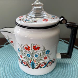 May include: A white enamel coffee percolator featuring a floral pattern in red, blue, and green. The pot has a black handle, a black rim, and a clear glass knob on the lid. The floral design is painted on the front and the lid of the vessel.