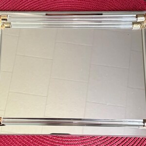 May include: A rectangular mirrored tray featuring a reflective glass surface. The tray is framed by four clear acrylic rods that run along each edge, secured at the corners by polished gold-tone metal connectors that provide a metallic accent.