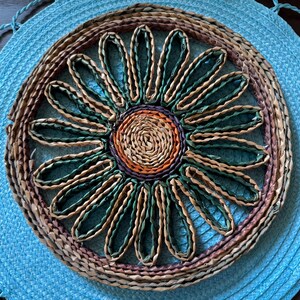 May include: A round, woven placemat with a sunflower design. The center is a spiral of natural fibers, surrounded by petals in shades of green, brown, and purple. The outer rim is a woven border of natural and brown fibers.