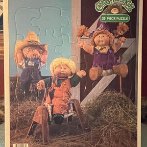May include: A 25-piece puzzle featuring three Cabbage Patch Kids dolls. The dolls are dressed in different outfits, including a cowboy hat, a purple dress, and a brown vest. The puzzle is set against a background of hay.