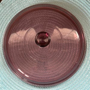 May include: A round, transparent, purple glass lid with a circular handle. The lid has concentric circles etched into the surface. The lid is sitting on a light blue woven placemat.
