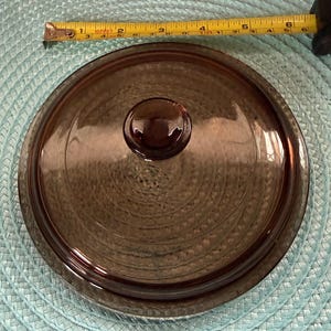 May include: A round, brown glass lid with a matching knob. The lid is approximately 6 inches in diameter. The lid is made of a transparent, heat-resistant glass, suitable for cookware. The lid is designed to fit a round pot or dish.