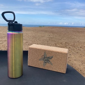 May include: A stainless steel water bottle with a rainbow iridescent finish and a black lid with a carrying handle. A cork yoga block with the text "Coastal Calm Yoga Co." is on a black yoga mat.