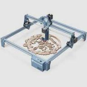 May include: A gray laser engraving machine with a wooden decorative piece in the center. The machine has a rectangular frame and a laser head that moves across the surface. The wooden piece has an ornate design with the words "Just Married" cut out.