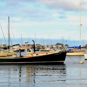 May include: A harbor scene with several sailboats and a dark green boat in the foreground. The boats are docked in calm water under a cloudy sky. A lighthouse is visible in the distance.