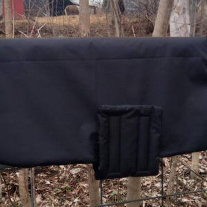 May include: A black dog coat with a quilted chest protector is draped over a metal fence. The coat has a rectangular shape with a curved edge and a small strap with a clip. The background shows trees and foliage.