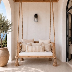 May include: A wooden porch swing suspended by thick jute ropes, featuring a white seat cushion and several beige throw pillows with varied textures. The frame has a cross-brace design on the sides, and the ropes end in decorative tassels.