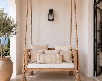 May include: A wooden porch swing suspended by thick jute ropes, featuring a white seat cushion and several beige throw pillows with varied textures. The frame has a cross-brace design on the sides, and the ropes end in decorative tassels.