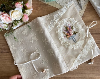 May include: Open, cream-colored book cover with floral embroidery and lace trim. The cover features a detailed embroidered bouquet within a lace oval, and a delicate floral pattern on the left side. A ribbon and wooden bead closure is visible.