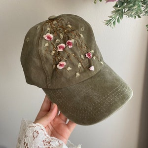 May include: A green baseball cap with embroidered pink flowers and vines. The cap has a curved brim and a structured crown.