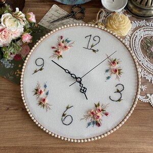 May include: A white embroidered clock with a pearl-like trim. The clock face features black Roman numerals and black hands. The embroidery includes pink and blue flowers.