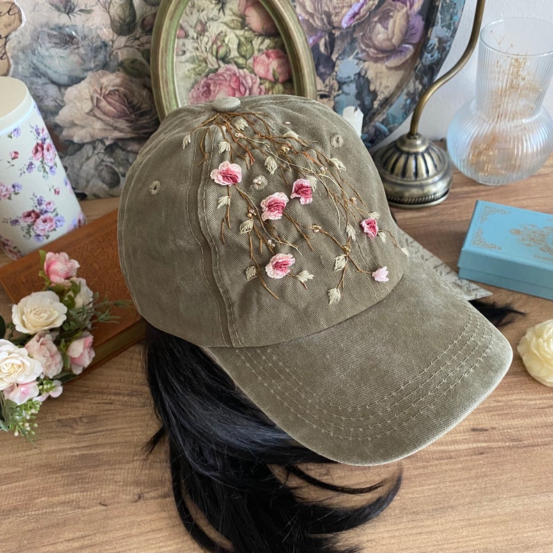 May include: A green baseball cap with pink and white embroidered flowers. The flowers are arranged in a cascading pattern on the front of the cap.