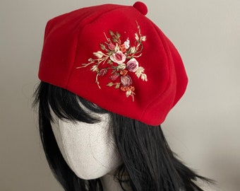 May include: A red beret with a floral embroidery design. The embroidery features pink, red, and white flowers with green leaves.
