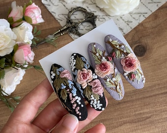 Puede incluir: Conjunto de tres pinzas para el pelo bordadas con flores. Las pinzas son de forma ovalada y presentan una variedad de flores rosas, blancas y azules con hojas verdes. Las pinzas están sobre un fondo blanco.
