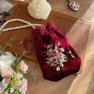 May include: A red velvet drawstring bag with a delicate lace trim and embroidered floral design. The bag is closed and sitting on a brown leather-bound book.
