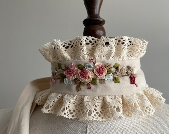 Puede incluir: Un collar de encaje blanco con una banda de flores bordadas en tonos rosas y verdes. El collar está en un maniquí blanco.