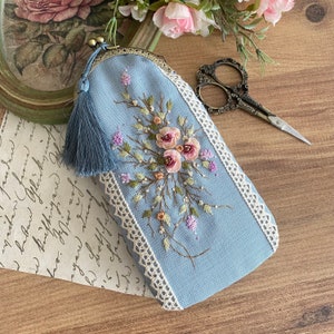 May include: A light blue fabric pouch with a white lace trim and embroidered flowers. The pouch has a tassel and is closed with a metal clasp.