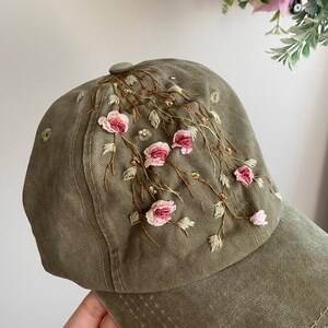 May include: A green baseball cap with embroidered pink flowers and vines. The cap has a curved brim and a strap at the back.