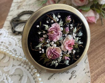 Puede incluir: Un compacto redondo de terciopelo negro con un marco de metal dorado. El compacto está decorado con un diseño floral cosido a mano en tonos de rosa y verde.
