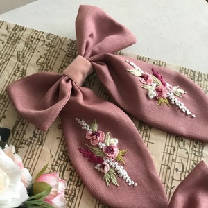 May include: A dusty rose fabric bow with two embroidered floral designs. The flowers are pink, white, and green.