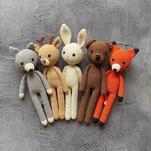 Può includere: Cinque animali di peluche all'uncinetto: un lupo grigio, un cervo marrone, un coniglietto bianco, un orso marrone e una volpe arancione. Gli animali sono tutti in piedi e hanno facce ricamate.