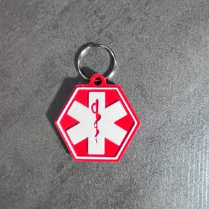 May include: A hexagonal medical alert keychain featuring a bright red background with a white Star of Life symbol and a central red staff of Asclepius. The emblem is attached to a circular silver metal split ring for secure attachment to keys.