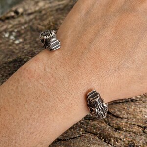 Könnte beinhalten: Ein silberfarbenes Armreif-Armband mit einem detaillierten, geformten Design. Das Armband hat zwei dekorative Enden mit einem strukturierten, mechanischen Aussehen. Das Armband wird am Handgelenk getragen und zeigt die komplizierte Metallarbeit.