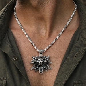 Op de afbeelding: Zilverkleurige ketting met een hanger met een gestileerde wolfskop. De hanger hangt aan een dikke, geweven ketting. De ketting wordt gedragen over een donkergroen overhemd.