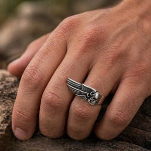 Könnte beinhalten: Ein silberner Ring mit einem Totenkopf und Flügeldesign. Der Ring wird an einem Finger getragen und hat ein detailliertes Design. Das Design des Rings umfasst einen Totenkopf mit Flügeln und besteht aus glänzendem Metall.