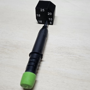 Pode incluir: Uma caneta preta e verde com uma ponta verde e um dado hexagonal preto com números brancos 5, 10, 15, 18, 20 e 25.