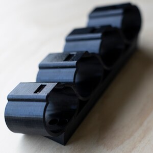 K&ouml;nnte beinhalten: Ein schwarzer, 3D-gedruckter Halter aus Kunststoff f&uuml;r vier zylindrische Gegenst&auml;nde. Der Halter ist so konzipiert, dass er die Gegenst&auml;nde sicher an ihrem Platz h&auml;lt.