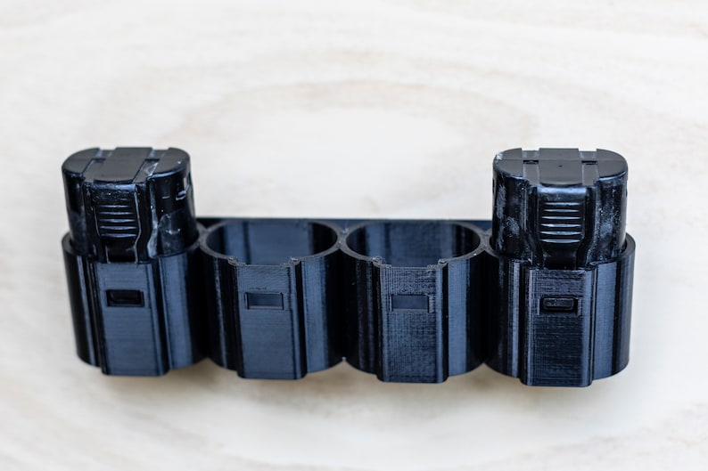 K&ouml;nnte beinhalten: Ein schwarzer Batteriehalter aus Kunststoff mit vier Steckpl&auml;tzen zum Aufbewahren von Batterien. Zwei Batterien befinden sich im Halter.