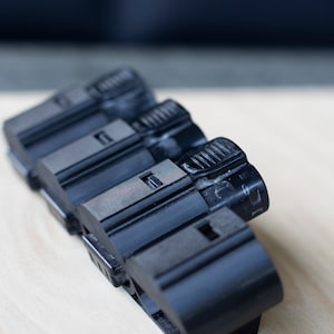 K&ouml;nnte beinhalten: Vier schwarze Batteriepacks aus Kunststoff, die auf einer hellen Holzoberfl&auml;che gestapelt sind. Die Batteriepacks haben eine strukturierte Oberfl&auml;che und eine kleine rechteckige &Ouml;ffnung oben.