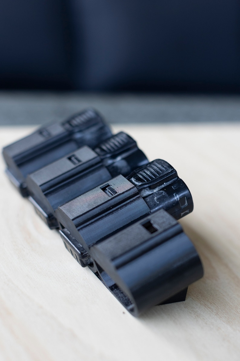 K&ouml;nnte beinhalten: Vier schwarze Batteriepacks aus Kunststoff, die auf einer hellen Holzoberfl&auml;che gestapelt sind. Die Batteriepacks haben eine strukturierte Oberfl&auml;che und eine kleine rechteckige &Ouml;ffnung oben.